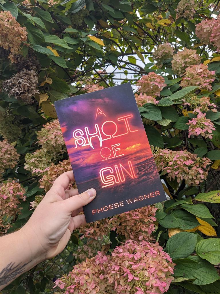 A white hand holds the book A Shot of Gin, which has a pink and purple front. In the background, pink hydrangea flowers match the colors of the book.
